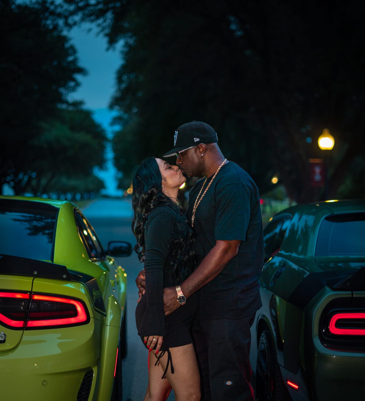 Couple kissing between two muscle cars on a tree-lined street at night