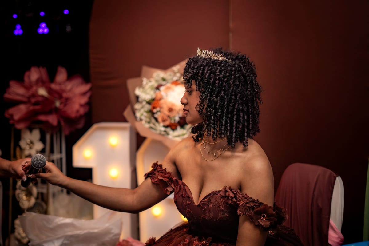 Sweet sixteen guest of honor in a tiara reaching for a microphone in front of her lit-up 16 sign