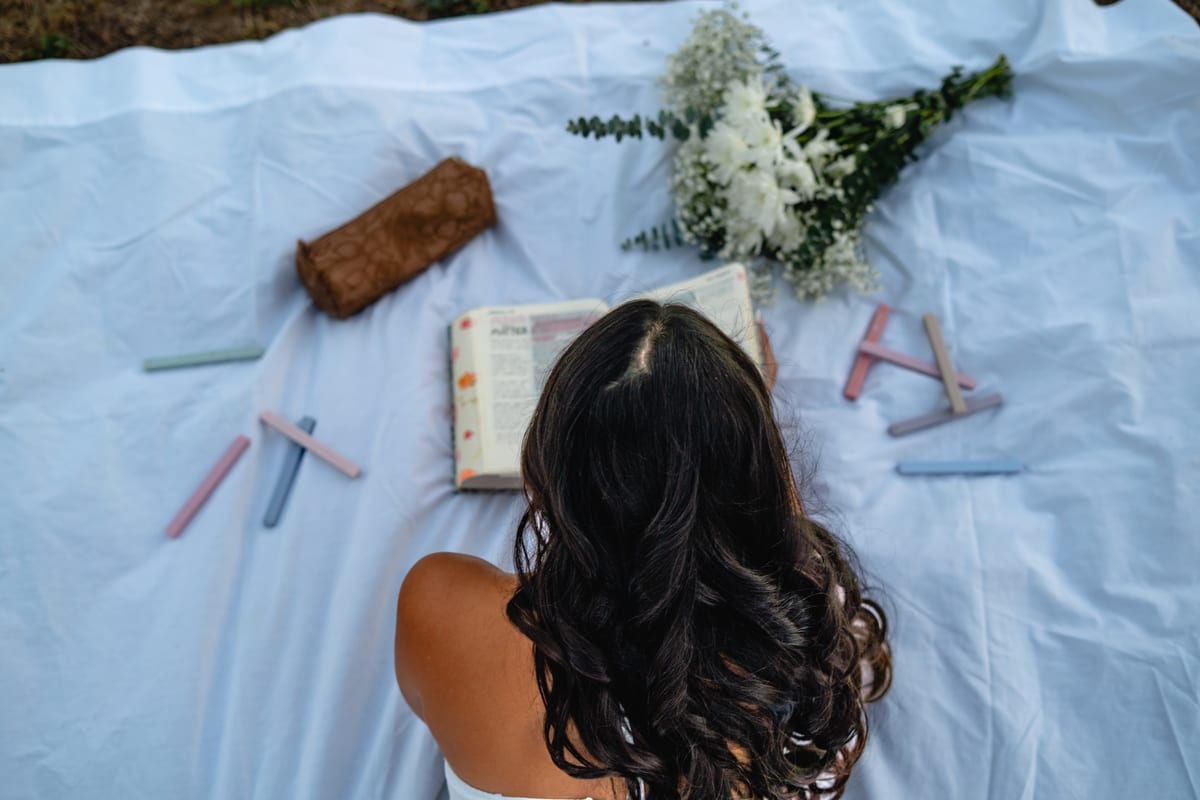 Overhead view of a young woman reading a Bible on a white blanket with flowers and chalk