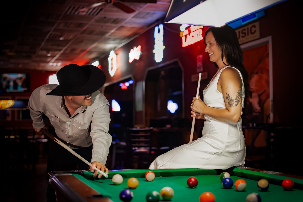 Man in a cowboy hat lining up a pool shot while a woman in a white dress sits on the table